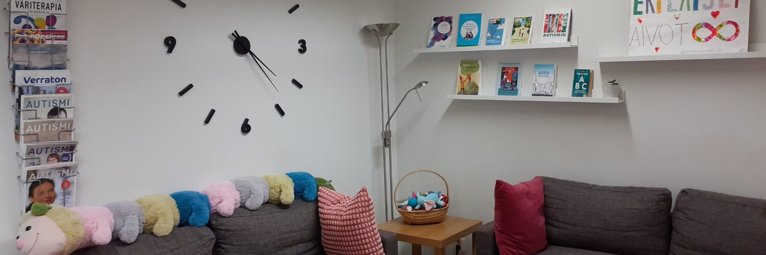 Kuvassa on toimisto, jossa näkyy kaksi sohvaa, joilla on tyynyjä ja pehmolelu, iso pöytä ja pienempi apupöytä sekä erilaisia kirjoja ja lehtiä. Seinällä on kello.