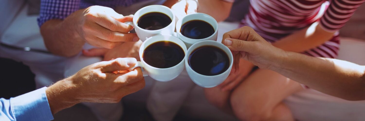 Neljän ihmisen kädet pitelevät kahvikuppeja keskellä. ihmisistä kahden vartalot näkyvät istumassa vaalealla sohvalla rinnasta alaspäin.