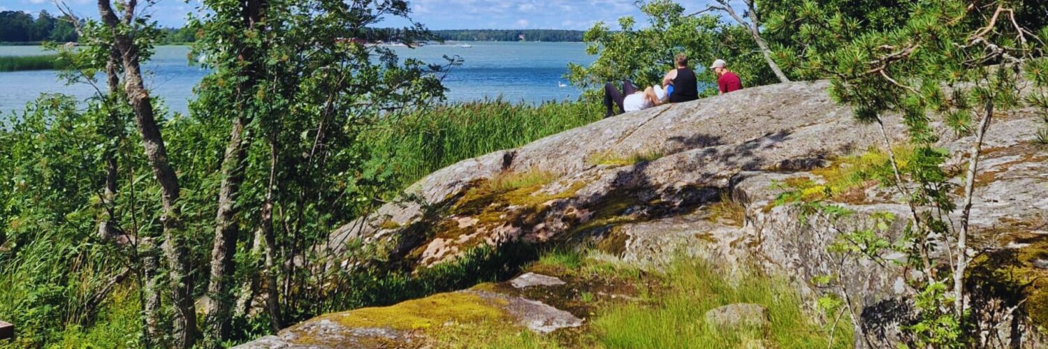 Kesäinen merenrantamaisema, jossa taustalla näkyy sinistä merta ja etualalla koivuja, puistikkoa ja rantakallio. Rantakalliolla istuu kolme henkilöä selät katsojaan päin. Etualalla vasemmalla on puinen penkki.