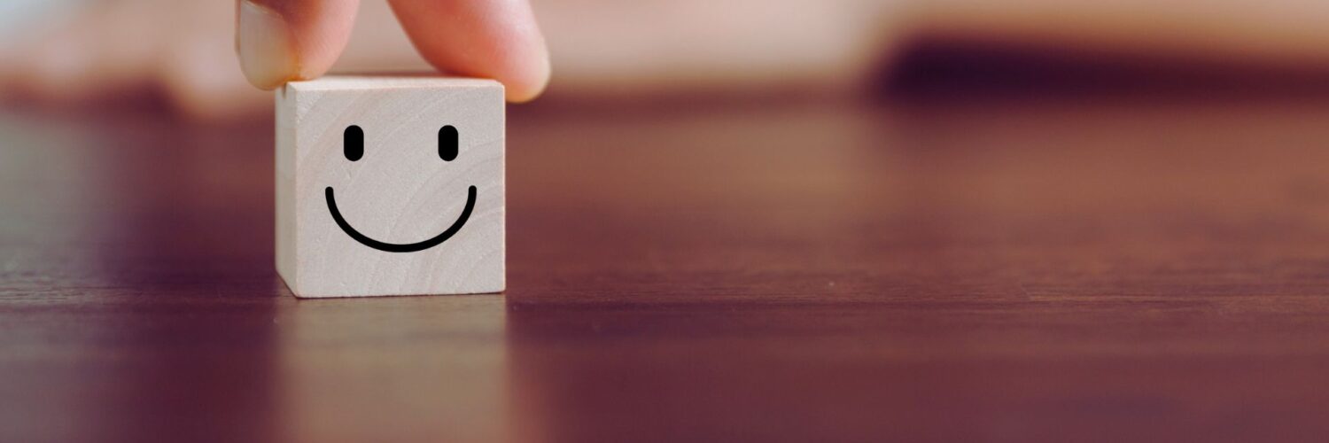 Puisella pöydällä pieni puinen kuutio, jonka kyljessä on hymyilevä hymynaama. Kuutiota pitelevät valkoihoisen ihmisen sormet.